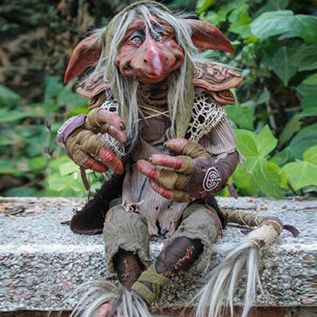 Gothic goblin sculpture with white beard and ragged clothes sitting on stone ledge in forest setting