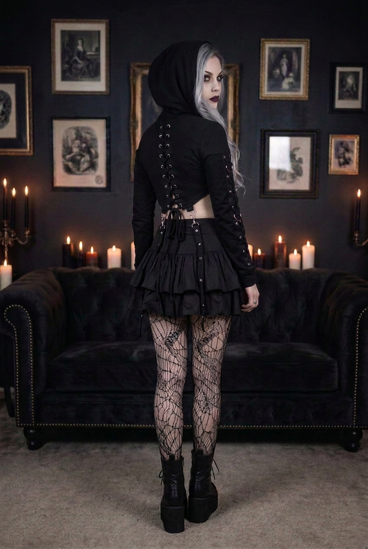 Goth model with silver-gray hair wearing black lace-up crop hoodie showing full back lace-up corset detail with O-ring hardware, layered ruffle mini skirt and spiderweb fishnet tights, back view in candlelit gothic interior — Shadow Realm Supply