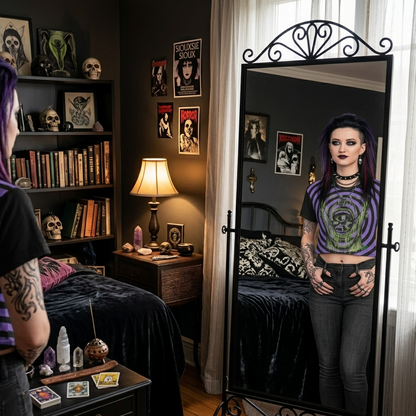 A person with purple hair and tattoos stands in a dark bedroom, looking at their reflection in a tall black metal freestanding mirror. They are wearing a purple and black striped graphic crop top with a green skull design and dark gray jeans.
