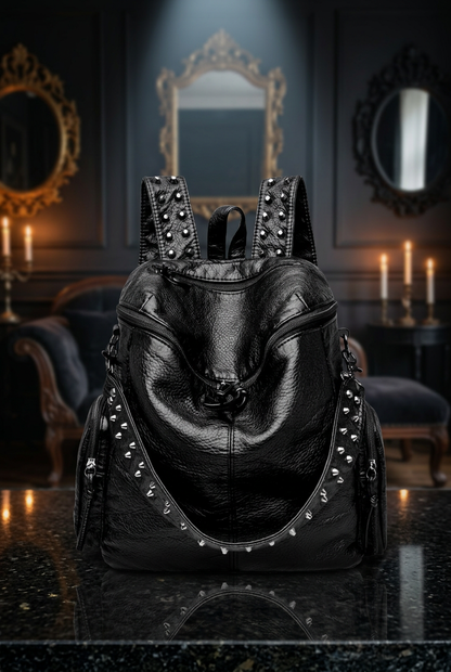 Gothic studded black vegan leather backpack front view with silver spike trim and chain detail, displayed on black marble surface in candlelit gothic interior with ornate gold mirrors — Shadow Realm Supply