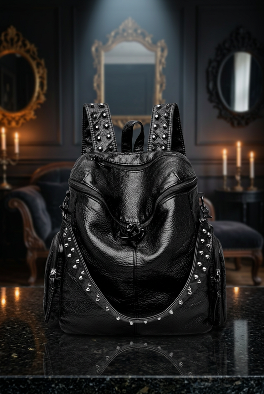 Gothic studded black vegan leather backpack front view with silver spike trim and chain detail, displayed on black marble surface in candlelit gothic interior with ornate gold mirrors — Shadow Realm Supply