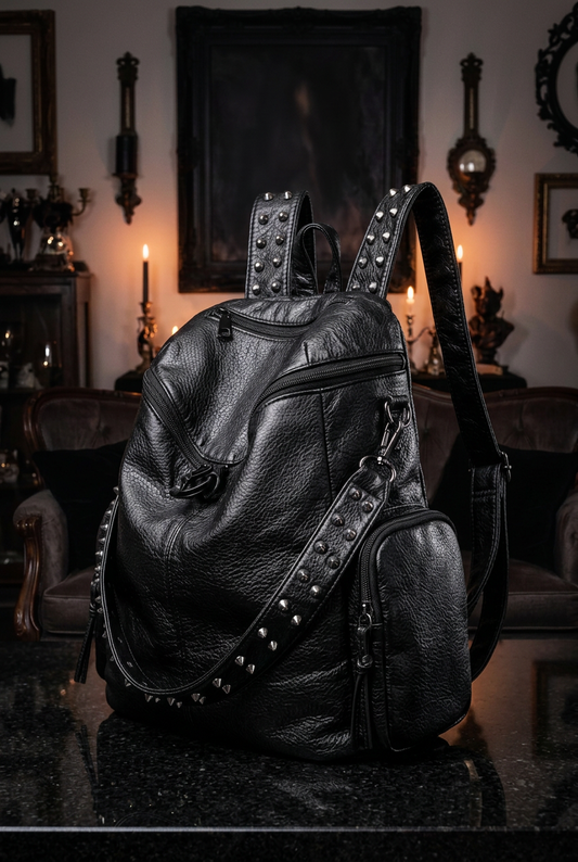 Gothic studded black vegan leather backpack three-quarter view showing detachable mini pouch and studded strap, displayed on black marble surface in moody candlelit gothic room — Shadow Realm Supply