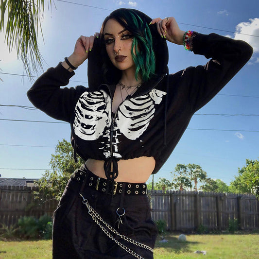 Alt model with black and green hair wearing black skeleton ribcage print zip-up crop hoodie with grommet belt and chain, hands pulling hood up, outdoor lifestyle shot — gothic punk alt fashion crop hoodie — Shadow Realm Supply