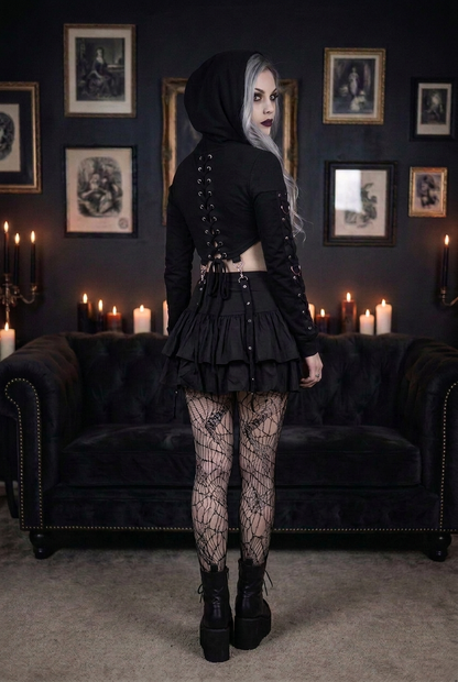 Goth model with silver-gray hair wearing black lace-up crop hoodie showing full back lace-up corset detail with O-ring hardware, layered ruffle mini skirt and spiderweb fishnet tights, back view in candlelit gothic interior — Shadow Realm Supply